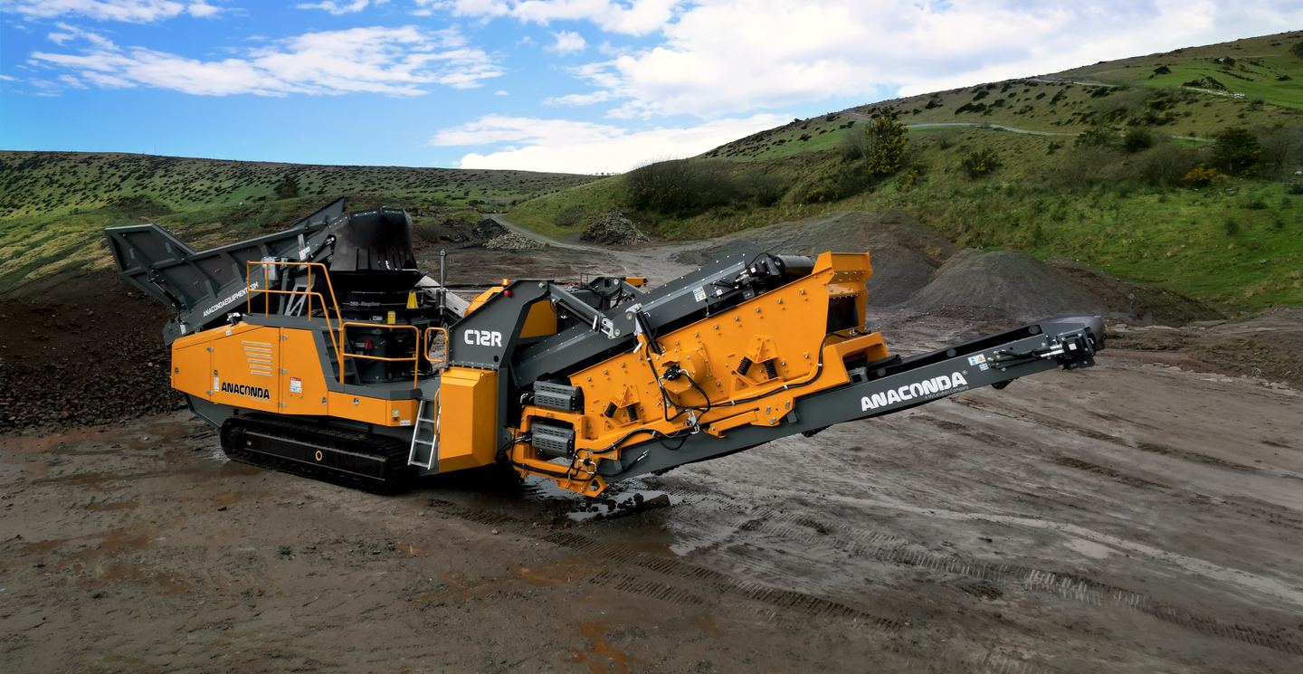 An orange Anaconda tracked screening plant working on a gravel site below green hills
