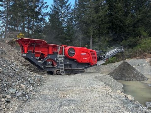 The 2023 Finlay J-1160 jaw crusher on a gravel job site among pine trees